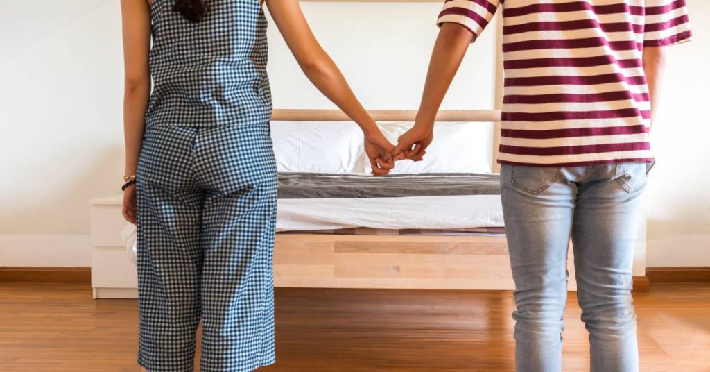 A man and woman hold hands while standing in front of a bed. The woman wears plaid, and the man wears a striped shirt.