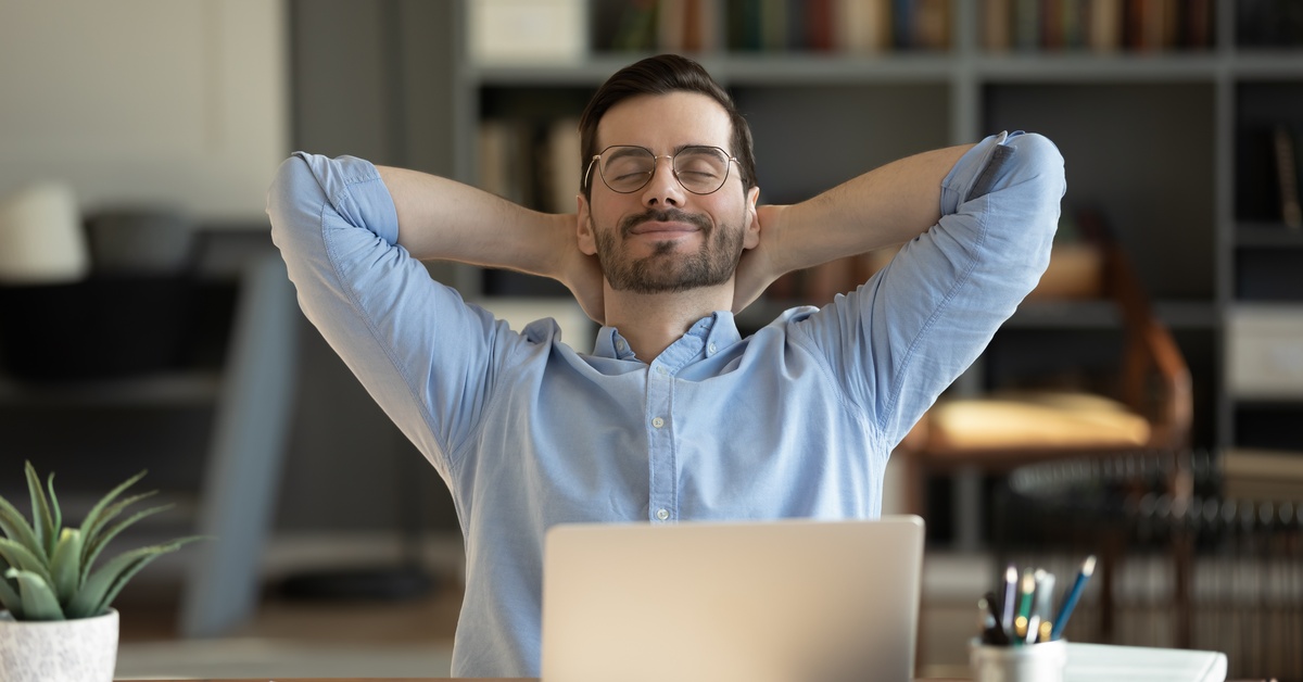 A man leaning back in his chair at his work desk, with his hands supporting his neck. He is smiling with his eyes closed.