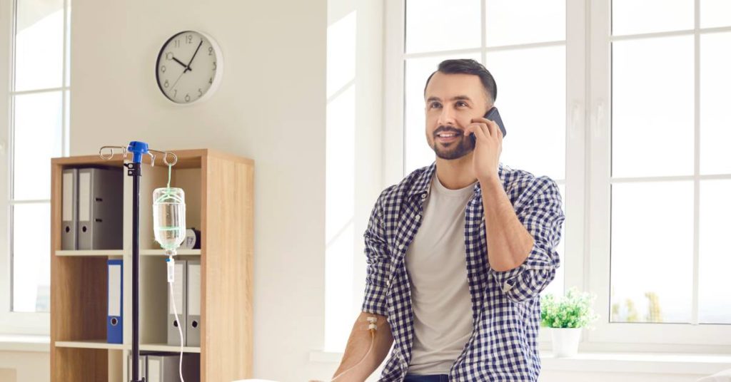 A man receives IV ozone treatment while sitting upright and talking on the phone. He wears a plaid shirt and jeans.