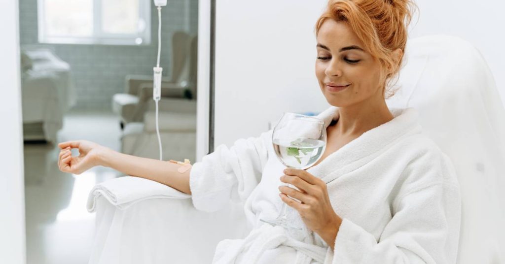 A woman receives IV ozone treatment in a plush chair while holding a glass of water. She smiles and wears a white robe.
