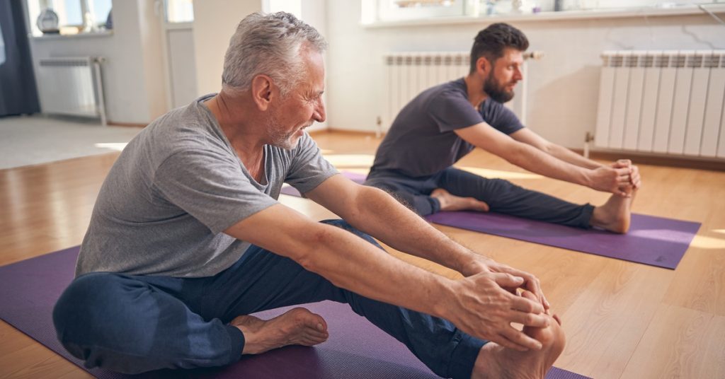 Two men practice yoga together in a studio. One man has gray hair and the other has brown hair. They both have purple mats.