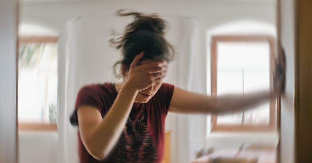 A woman with messy brown hair holds her head and grasps onto a wall. Her background is slightly blurred.