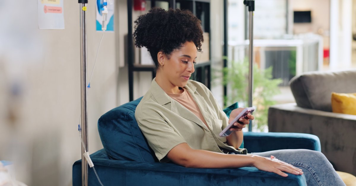 A woman sitting in a chair with an IV attached to her arm. She is looking at her phone and there is another chair beside her.