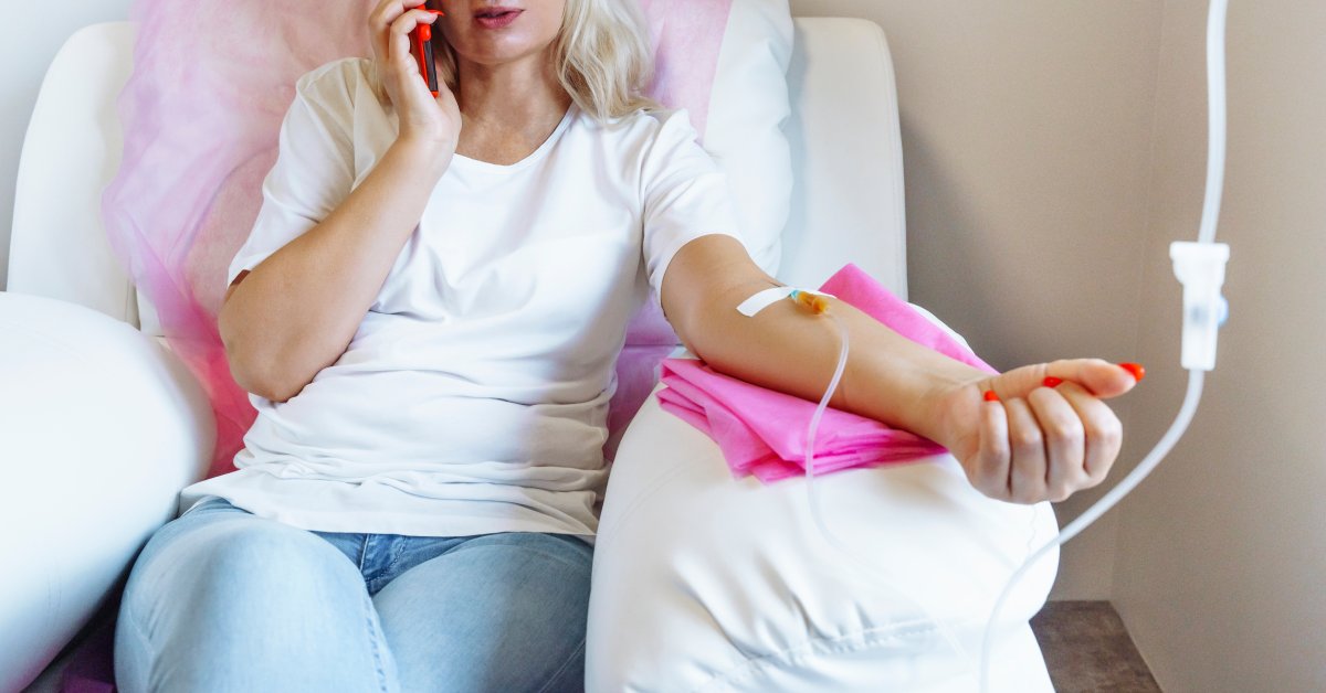 A woman is sitting on a chair talking on her phone. She has an IV in her arm and her arm is resting on a pink cloth.
