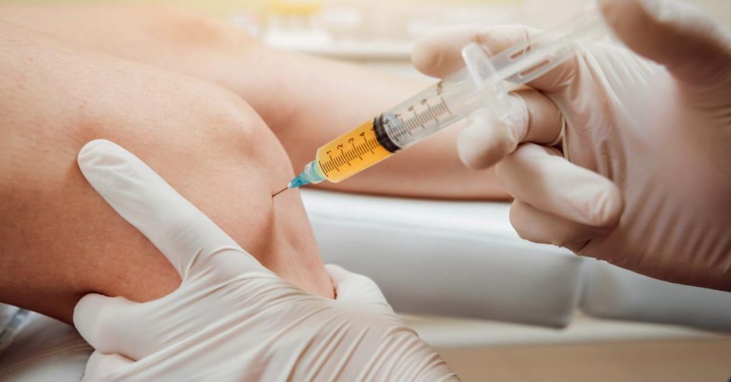 A close-up of a knee receiving a PRP injection. The administrator wears white gloves and grips the knee.