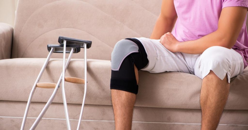 A close-up of a man sitting on a couch with a black-and-gray knee brace. A pair of crutches sits beside him.