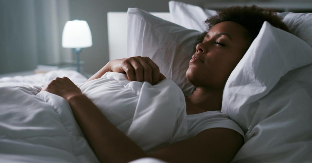 A close-up of a Black woman sleeping in bed. She props her head up on two pillows, and she crosses her arms on her comforter.