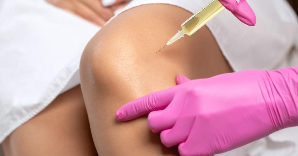 A close-up of a practitioner wearing pink latex gloves and holding a fluid-filled needle to a patient's knee.