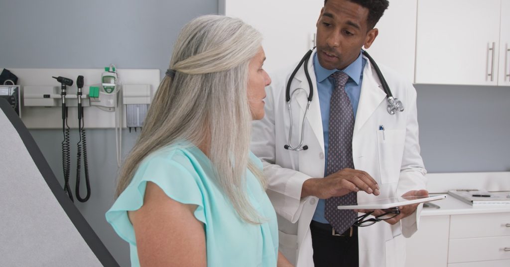 A woman is sitting in a room, there is a doctor standing next to her. The doctor is holding a clipboard.