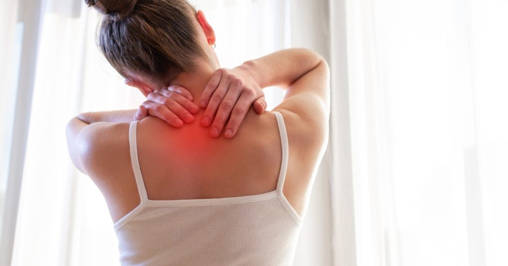 A woman facing a white curtain presses both of her hands against the back of her neck.