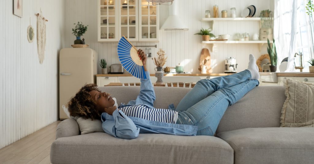 A woman is lying down on a grey couch and fanning her face with a blue folding hand fan.