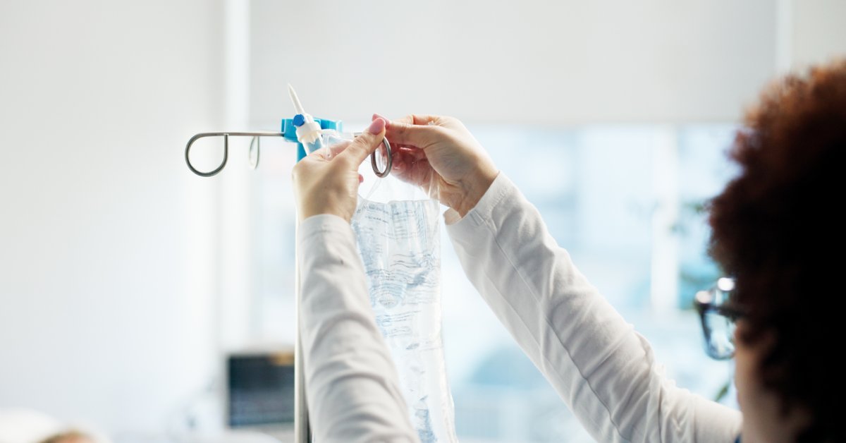 A person wearing a white long-sleeved shirt hangs an IV bag full of a clear liquid on an IV pole.