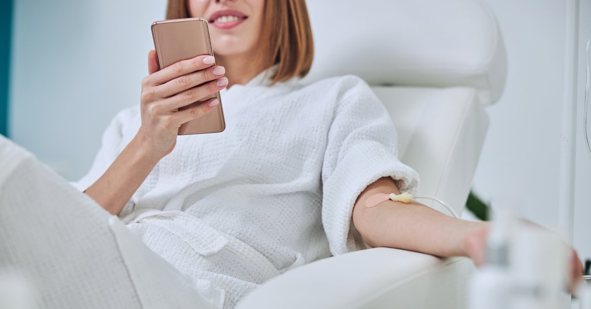 A woman sitting in a white armchair is smiling and looking at a mobile phone while she receives IV therapy.