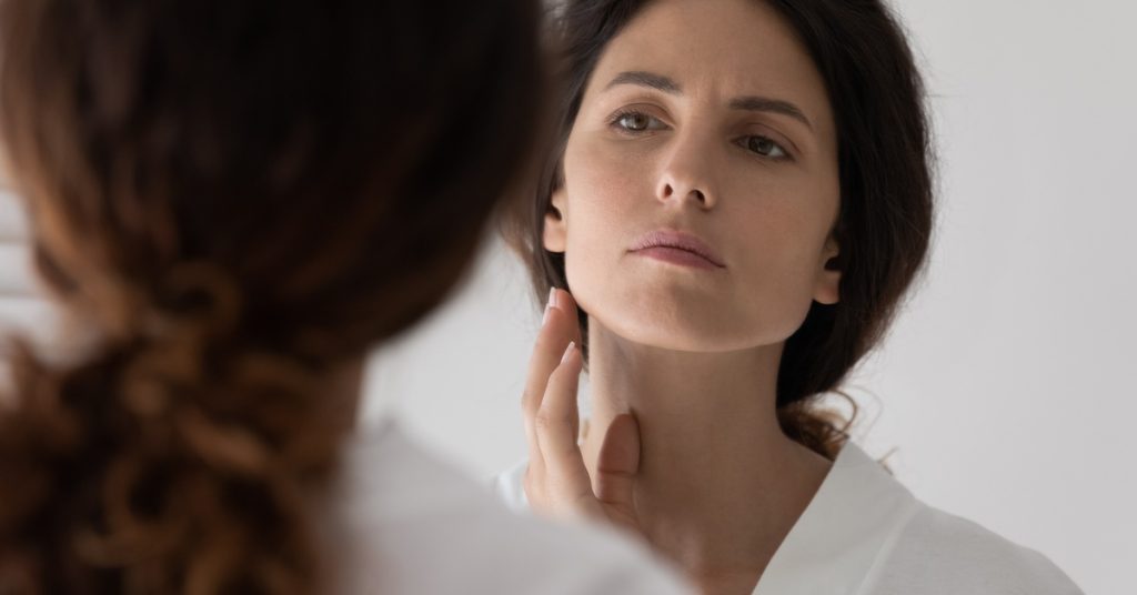 A woman with long brown hair is wearing a white robe and furrows her eyebrows while inspecting her face in a mirror.