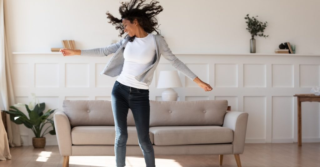 A happy woman with long brown curly hair is dancing in her living room.