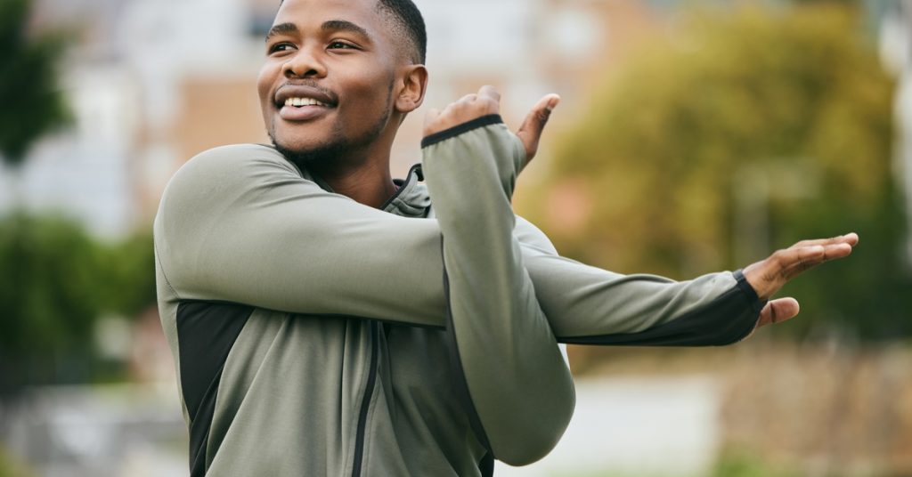A smiling man in an outdoor setting is performing a cross arm stretch to loosen up his shoulder muscles.