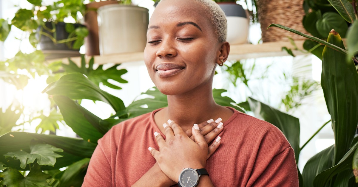 A relaxed woman wearing a pale red shirt and a black wristwatch closes her eyes and smiles while she rests her hands over her heart and sits in a room full of houseplants.