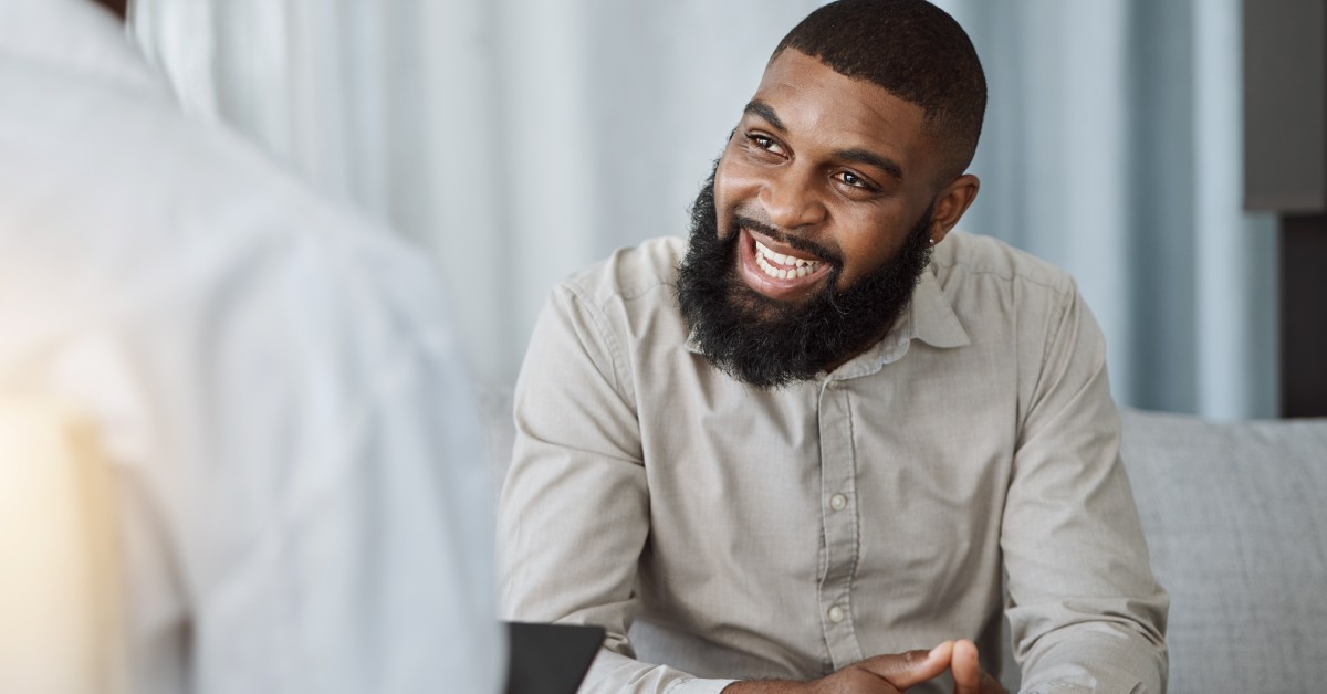A smiling man wearing a cream color button-up shirt sits on a grey couch and laces his fingers together while talking to a therapist.