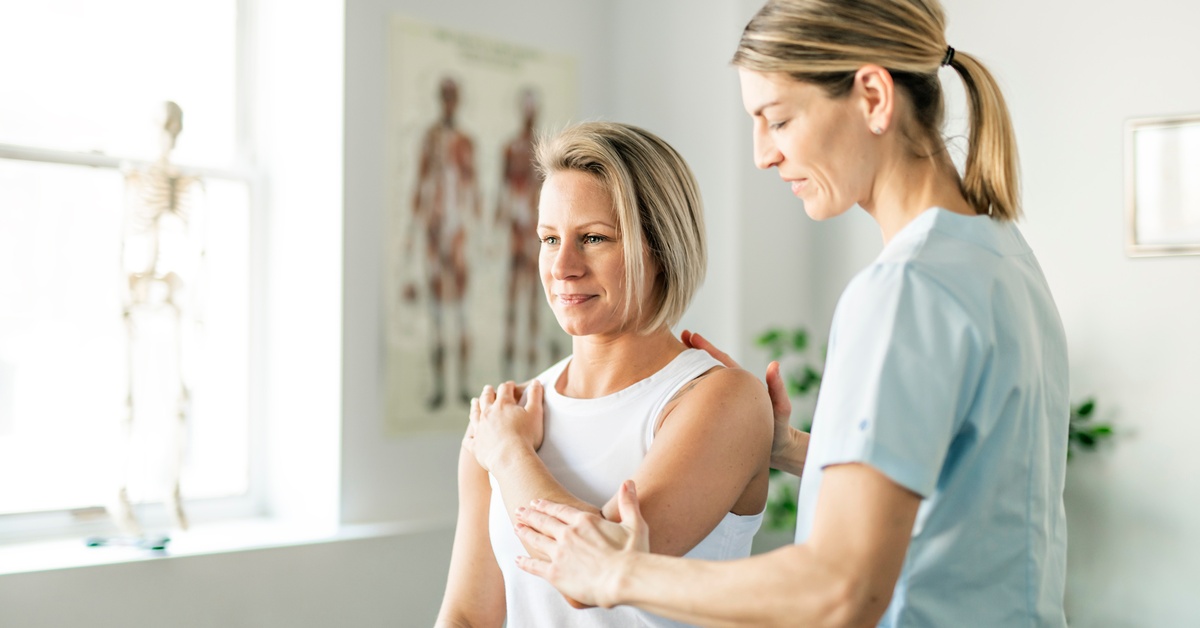 A woman crosses one arm over her chest to her shoulder while a rehabilitation nurse guides her in an exam room.