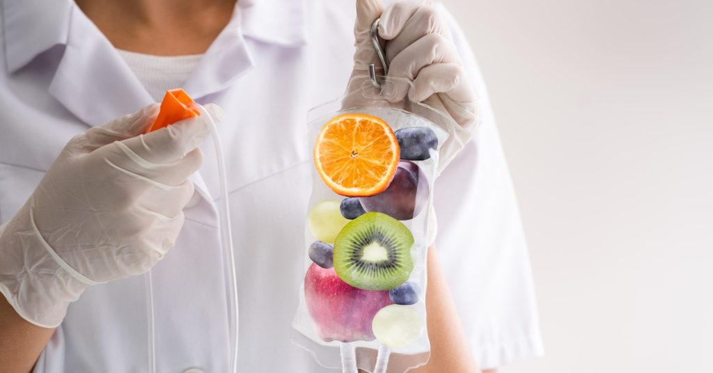 A nurse in white holds a clear IV bag containing fruits that symbolize essential nutrients for wellness.